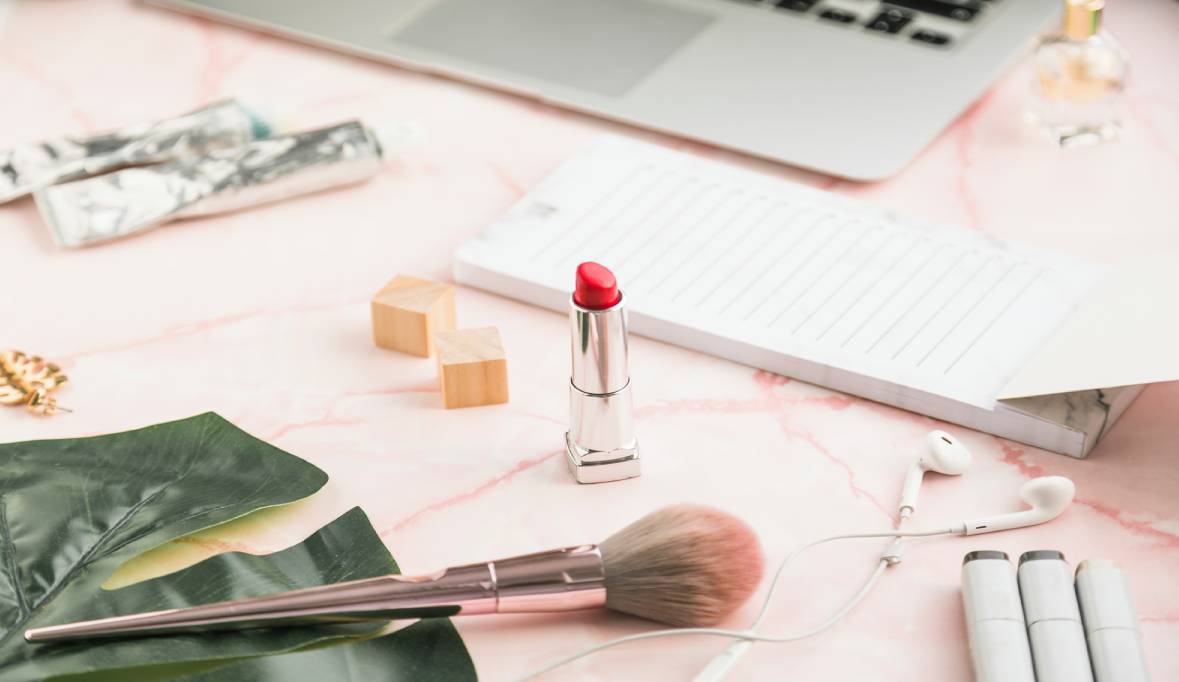 A stylish workspace with pink marble surface, featuring red lipstick and makeup brush