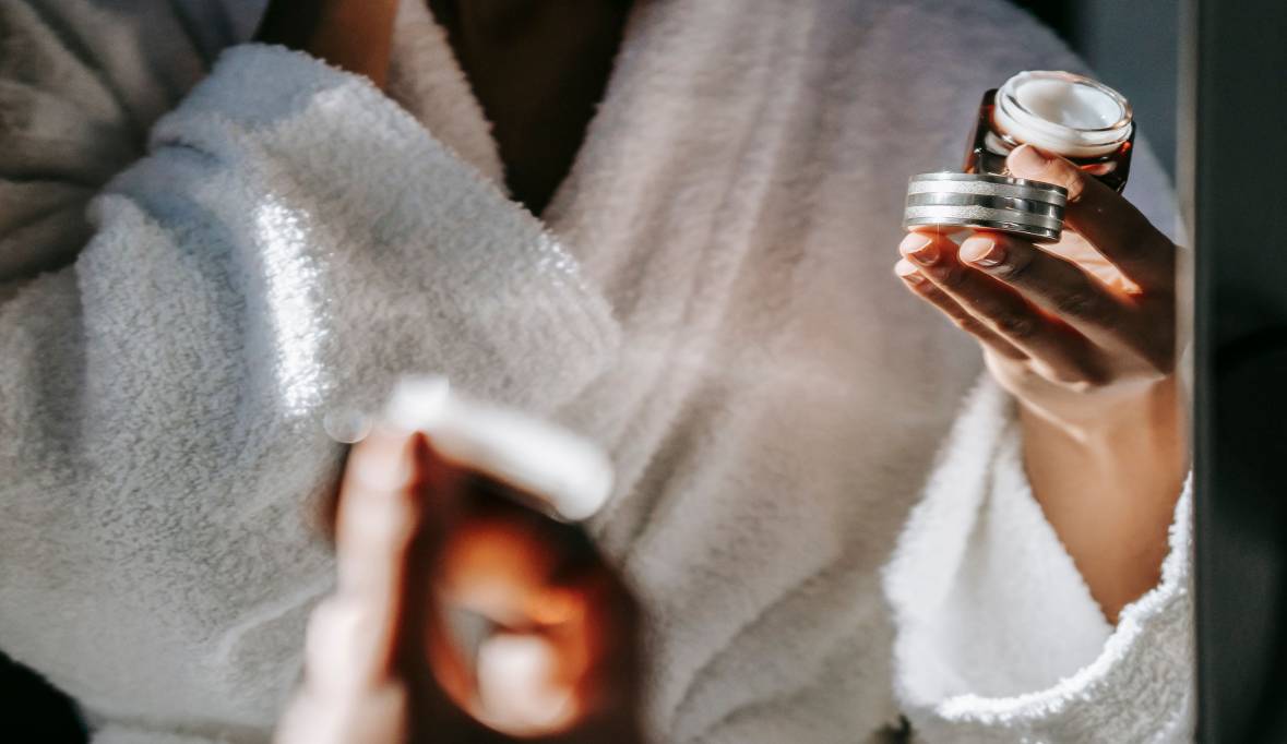A woman in a bathrobe holds a container of cream, preparing for her skincare routine