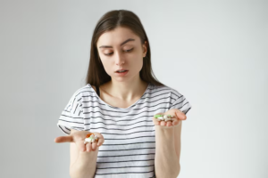 Isolated portrait doubtful pensive young european woman posing with open hands front her holding bunch multicolored pills her palms choosing antibiotics vitamins